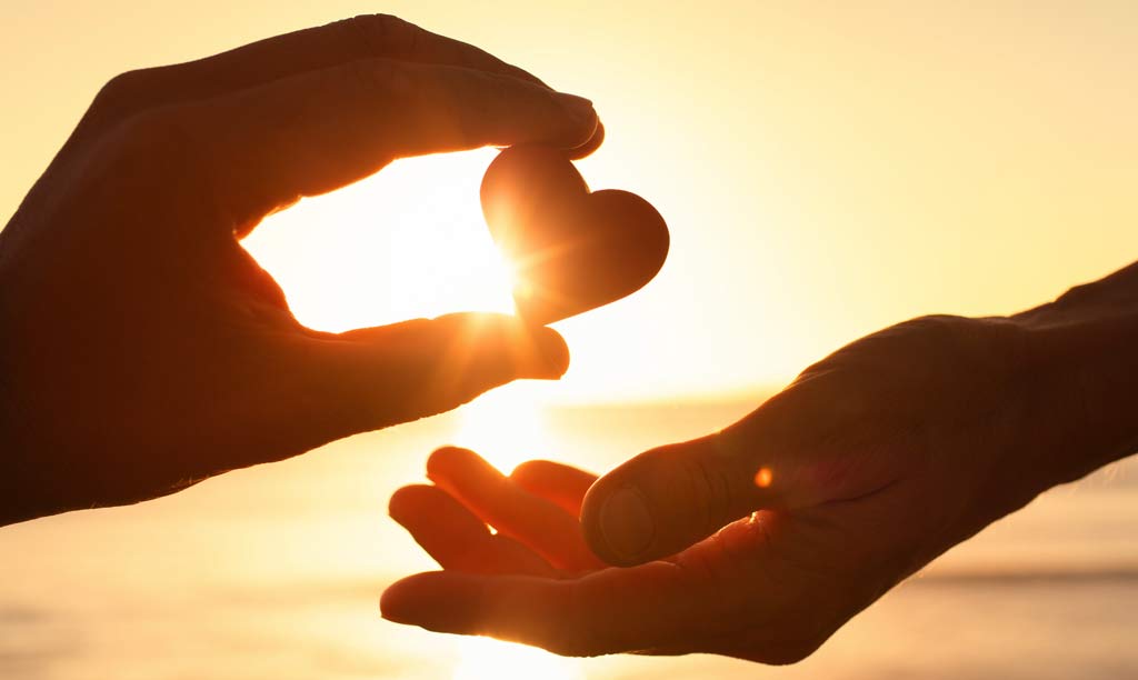 A hand holding a heart shaped stone up into the sunset